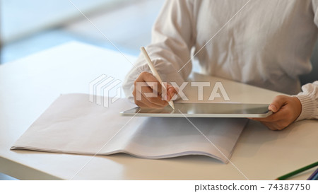 Cropped image of beautiful woman hands holding a stylus pen and computer tablet while drawing/sketching at the working desk over comfortable living room as background. Cropped image of beautiful woman hands holding a stylus pen and computer tablet while drawing/sketching at the working desk over comfortable living room as background. 74387750