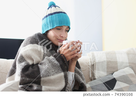 Chilled woman on couch in blanket and hat holds hot cup Chilled woman on couch in blanket and hat holds hot cup 74390108