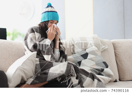 chilled woman on sofa in blanket and hat holds handkerchief 74390116