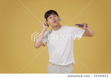 portrait of happy joyful cheerful young asian man wearing headphones listening to music on smartphone and dancing isolated on yellow background portrait of happy joyful cheerful young asian man wearing headphones listening to music on smartphone and dancing isolated on yellow background 74394915