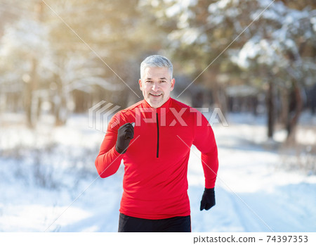 Cross country running in winter. Happy fit senior man jogging in snowy forest on sunny morning 74397353
