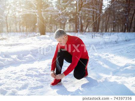 Mature jogger in warm sportswear tying shoelaces on snowy road at winter park Mature jogger in warm sportswear tying shoelaces on snowy road at winter park 74397356