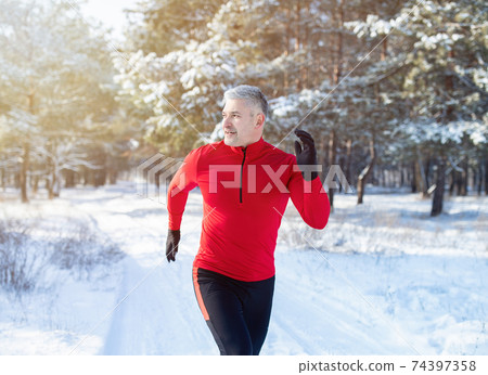 Cold weather running concept. Fit mature man in sports clothes jogging at snowy winter park. Seasonal workout Cold weather running concept. Fit mature man in sports clothes jogging at snowy winter park. Seasonal workout 74397358