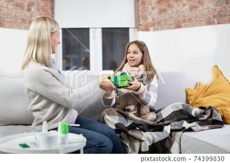 Portrait of happy surprised sick teen girl, smiling to her positive loving mother, presenting box gift to her daughter, supporting her while sitting together on cozy sofa at home 74399830