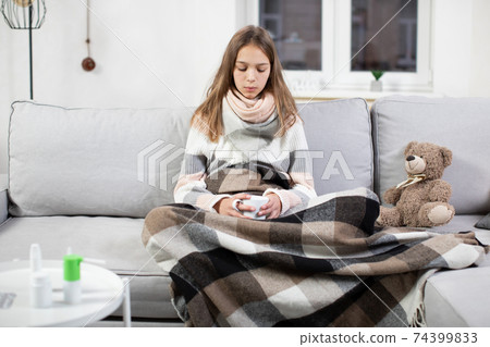 Cold and flu season. Sick teenage girl with high fever and sore throat, holding cup of hot tea and feeling bad, sitting at home on the sofa under warm blanket 74399833
