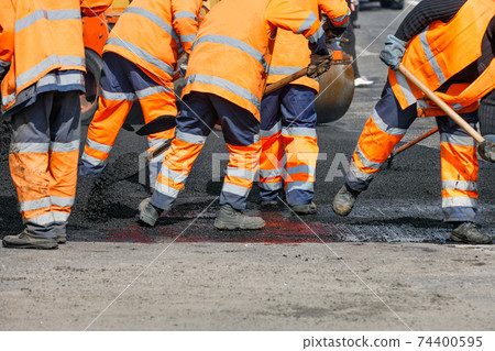 A team of road workers in orange reflective uniforms use shovels to scatter some of the asphalt to repair a section of the road on a clear day. 74400595