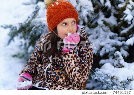 child girl sitting on a sled and tasting the snow in a winter forest, bright snowy fir trees, beautiful nature 74401157