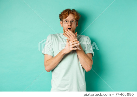 Shocked redhead man in glasses gasping startled, covering mouth and staring at camera scared, standing over turquoise background Shocked redhead man in glasses gasping startled, covering mouth and staring at camera scared, standing over turquoise background 74403090