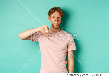 Disappointed redhead man showing thumbs-down and tongue, epxress dislike and disapproval, standing over turquoise background Disappointed redhead man showing thumbs-down and tongue, epxress dislike and disapproval, standing over turquoise background 74403583