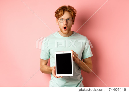 Excited young man with red hair and beard, checking out online promotion, showing digital tablet screen and staring at camera amazed, standing over pink background Excited young man with red hair and beard, checking out online promotion, showing digital tablet screen and staring at camera amazed, standing over pink background 74404446