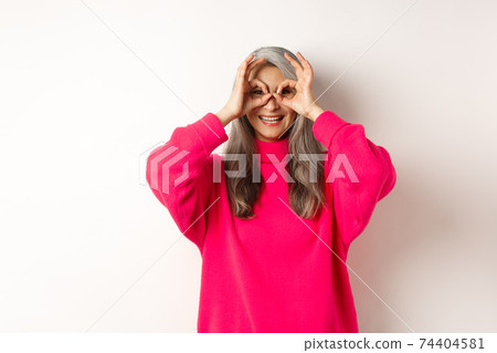 Happy and funny asian senior woman making hand binoculars, looking through fingers and smiling, standing in pink sweater over white background 74404581