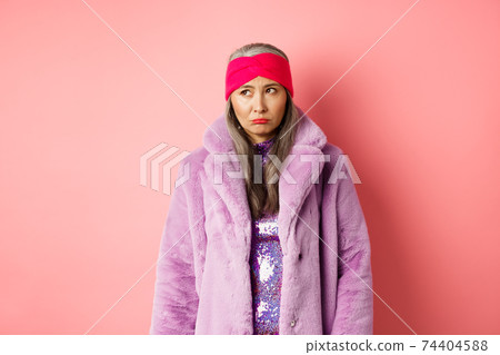 Sad and indecisive asian senior woman in fashionable faux fur coat looking left and sulking upset, standing against pink background 74404588
