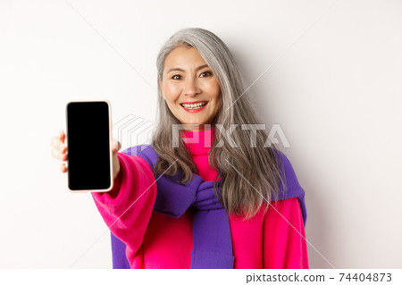 Online shopping. Close up of stylish asian senior woman extending hand with mobile phone, showing blank smartphone screen and smiling, standing over white background 74404873