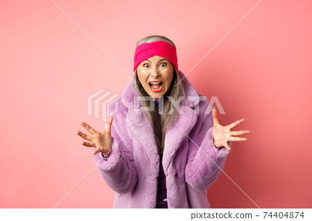 Angry asian senior woman in stylish purple faux fur coat, shaking hands and screaming at camera outraged, shouting at you, standing over pink background 74404874
