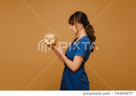 Cute young doctor girl holding a human skull, brown background behind the doctor Cute young doctor girl holding a human skull, brown background behind the doctor 74404977