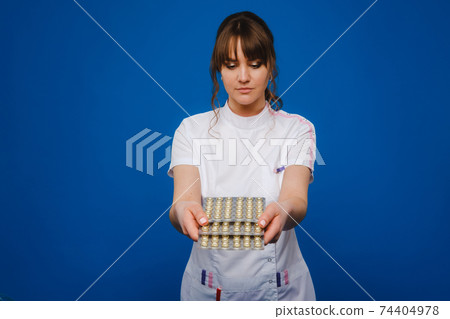 The concept of health care. A young brunette doctor in a white coat on a blue background shows plates with capsules to take. The concept of health care. A young brunette doctor in a white coat on a blue background shows plates with capsules to take. 74404978