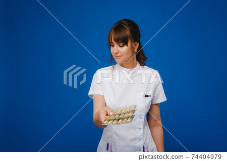The concept of health care. A young brunette doctor in a white coat on a blue background shows plates with capsules to take. The concept of health care. A young brunette doctor in a white coat on a blue background shows plates with capsules to take. 74404979