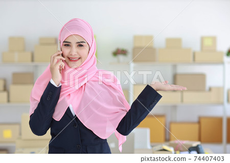 Religious asian muslim woman in blue suit and pink shaft on head standing and using mobile phone with confidence and showing something on hand with business package SME box delivery background. Religious asian muslim woman in blue suit and pink shaft on head standing and using mobile phone with confidence and showing something on hand with business package SME box delivery background. 74407403