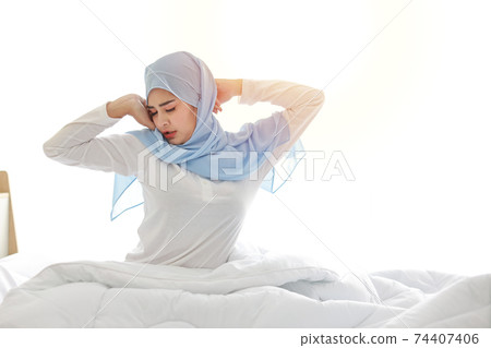 Beautiful asian muslim woman stretching her arms after getting up in the morning on bed at sunrise in bedroom. Cute young girl with blue hijab standing and relaxing with happy and smiling face. 74407406