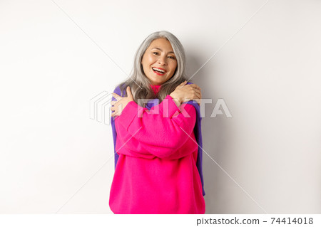 Valentines day and holidays concept. Lovely asian senior woman in pink sweater hugging herself with eyes closed, smiling, standing over white background 74414018