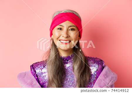 Close-up of trendy asian senior woman with grey hair and stylish disco dress, smiling happy at camera, standing over pink background 74414912