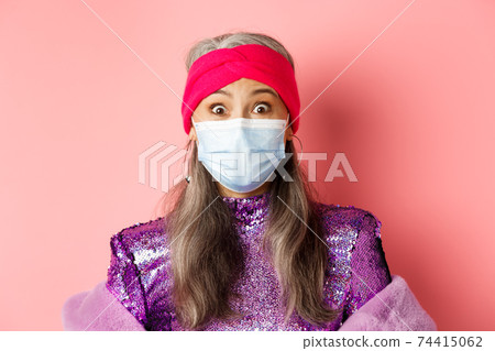 Covid-19, virus and social distancing concept. Close up of fashionable elderly woman in medical mask and headband staring surprised at camera, pink background 74415062