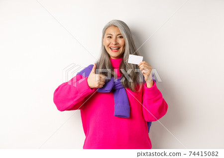 Shopping concept. Smiling asian middle-aged woman with grey hair showing plastic credit card and thumbs-up, recommending bank promotion, white background 74415082