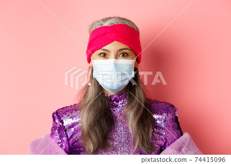 Covid-19, virus and social distancing concept. Close-up of stylish asian senior woman in medical mask and shiny dress looking at camera, standing over pink background 74415096