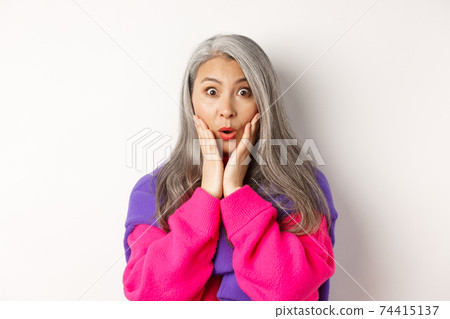 Close up of fashionable asian senior woman with grey hair looking surprised, saying wow and staring at camera amazed, standing over white background 74415137
