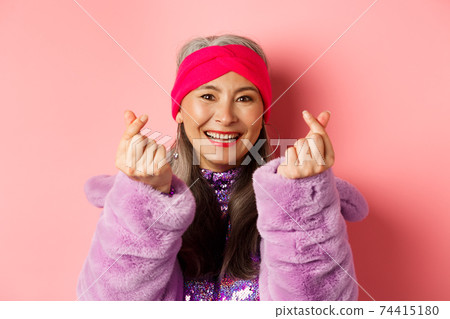 Fashion. Beautiful and fashionable asian senior woman smiling, showing korean heart gesture and looking happy, standing over pink background Fashion. Beautiful and fashionable asian senior woman smiling, showing korean heart gesture and looking happy, standing over pink background 74415180