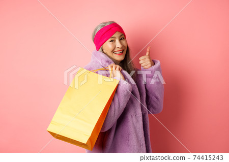 Stylish asian senior woman going shopping, carry paper bag on shoulder and showing thumb-up, recommending store discounts, pink background 74415243