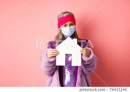 Covid-19 and real estate concept. Senior asian woman in stylish outfit and medical mask showing paper house model, searching apartment, standing over pink background 74415244
