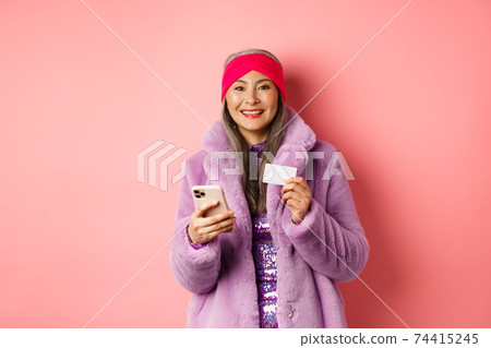 Online shopping and fashion concept. Smiling middle-aged woman in stylish fur coat using mobile phone and plastic credit card, standing happy on pink background 74415245