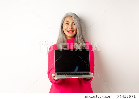 E-commerce concept. Beautiful asian senior businesswoman showing blank laptop screen and smiling, standing in pink sweater over white background E-commerce concept. Beautiful asian senior businesswoman showing blank laptop screen and smiling, standing in pink sweater over white background 74415671