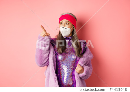 Covid and fashion concept. Impressed asian elderly woman checking out promo offer, wearing respirator from coronavirus, pointing and looking left, pink background Covid and fashion concept. Impressed asian elderly woman checking out promo offer, wearing respirator from coronavirus, pointing and looking left, pink background 74415948