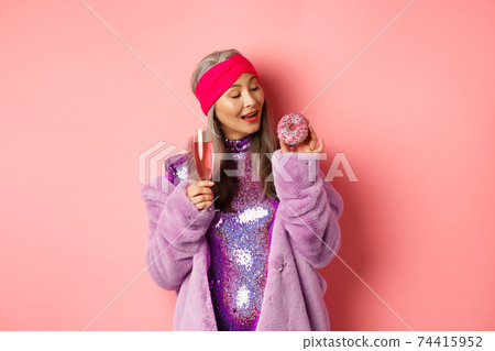 Fancy senior woman having fun, eating donut and drinking pink champagne, standing in purple faux fur coat and glittering dress, studio background Fancy senior woman having fun, eating donut and drinking pink champagne, standing in purple faux fur coat and glittering dress, studio background 74415952