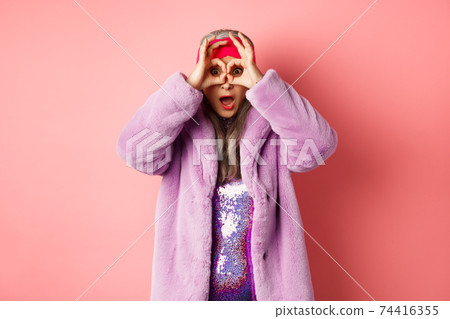 Fashion and shopping. Amazed asian senior woman checking out awesome valentines day promotion, looking through hands binoculars and gasping impressed, pink background 74416355
