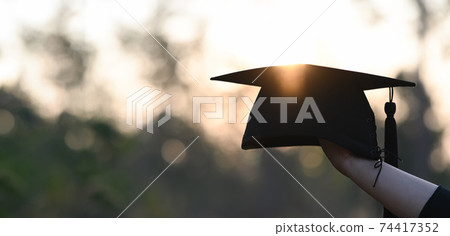 Cropped image of university student hand holding a graduation hat in hand over outdoors with sunset as background. Cropped image of university student hand holding a graduation hat in hand over outdoors with sunset as background. 74417352