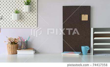 A white student desk that surrounded by bookshelf, coffee cup, diary, notebook, stack of books, pencil holder, flowers in wicker basket, potted plant and post it. 74417358