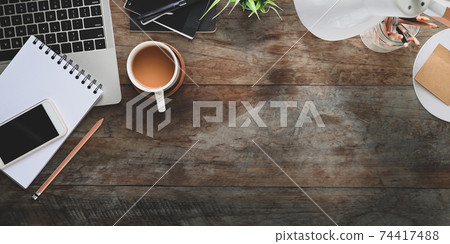 Top view of rustic table office desk with coffee cup putting surrounded computer laptop, note, pencils, pencil holder, potted plant , lamp, pen and smartphone. 74417488
