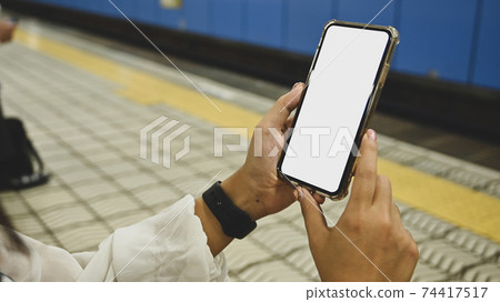Cropped image of attractive woman's hands holding a black smartphone with white blank screen over empty railway platform as background. Cropped image of attractive woman's hands holding a black smartphone with white blank screen over empty railway platform as background. 74417517