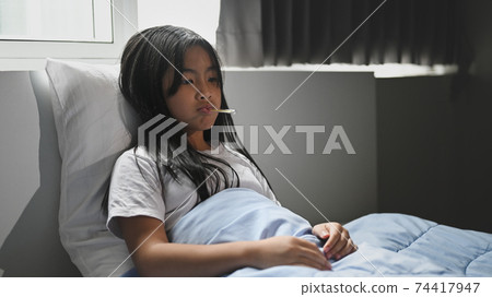 A little sick girl covered in a blanket is lying on the bed and measuring the temperature. A little sick girl covered in a blanket is lying on the bed and measuring the temperature. 74417947