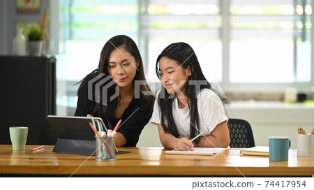 A young mother sits and doing homework together with her daughter at the wooden student desk. 74417954