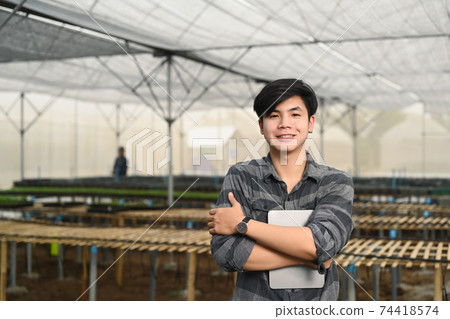 Smart young asian farmer man holding digital tablet while standing in greenhouse filled with organic vegetables. 74418574