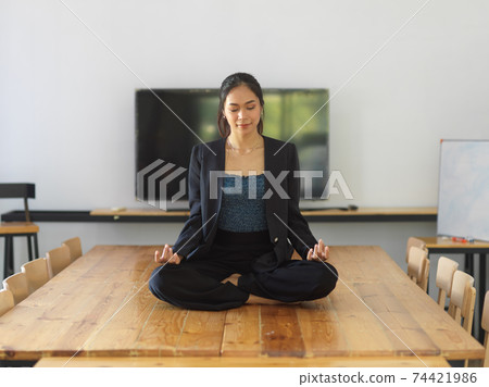 Portrait of female worker sitting in yoga position and meditating on the table Portrait of female worker sitting in yoga position and meditating on the table 74421986