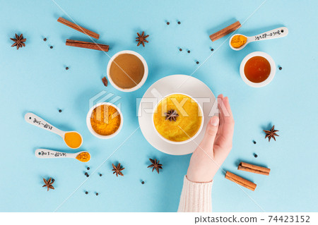 Turmeric golden milk latte in woman hand with spices, cinnamon and honey on blue background. Healthy traditional indian drink. 74423152