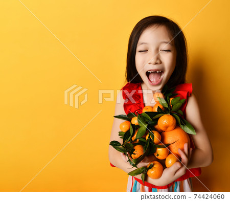 Asian child in red blouse. Looking wondered, holding an armful of tangerines and oranges, posing on orange background. Close up Asian child in red blouse. Looking wondered, holding an armful of tangerines and oranges, posing on orange background. Close up 74424060