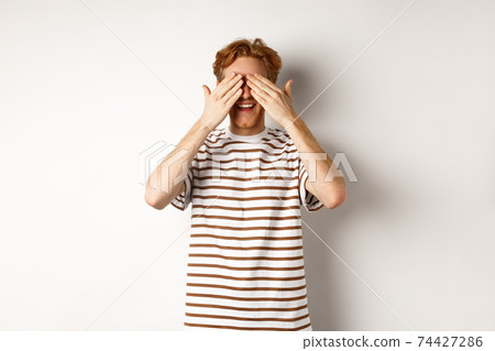 Smiling man cover eyes and waiting for surprise, standing in t-shirt against white background Smiling man cover eyes and waiting for surprise, standing in t-shirt against white background 74427286