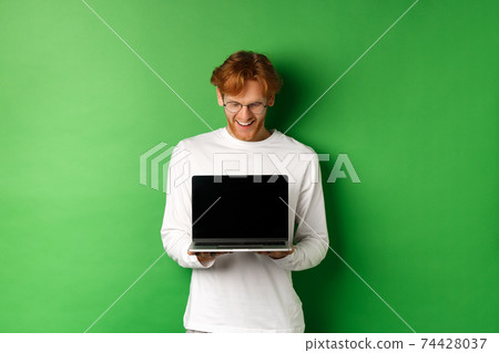 Happy young man with red hair, showing blank laptop screen and looking pleased, smiling while staring at computer, green background Happy young man with red hair, showing blank laptop screen and looking pleased, smiling while staring at computer, green background 74428037