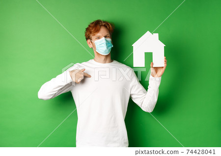 Real estate and covid-19 pandemic concept. Smiling redhead man in face mask pointing at paper house cutout, showing broker agency, standing over green background Real estate and covid-19 pandemic concept. Smiling redhead man in face mask pointing at paper house cutout, showing broker agency, standing over green background 74428041